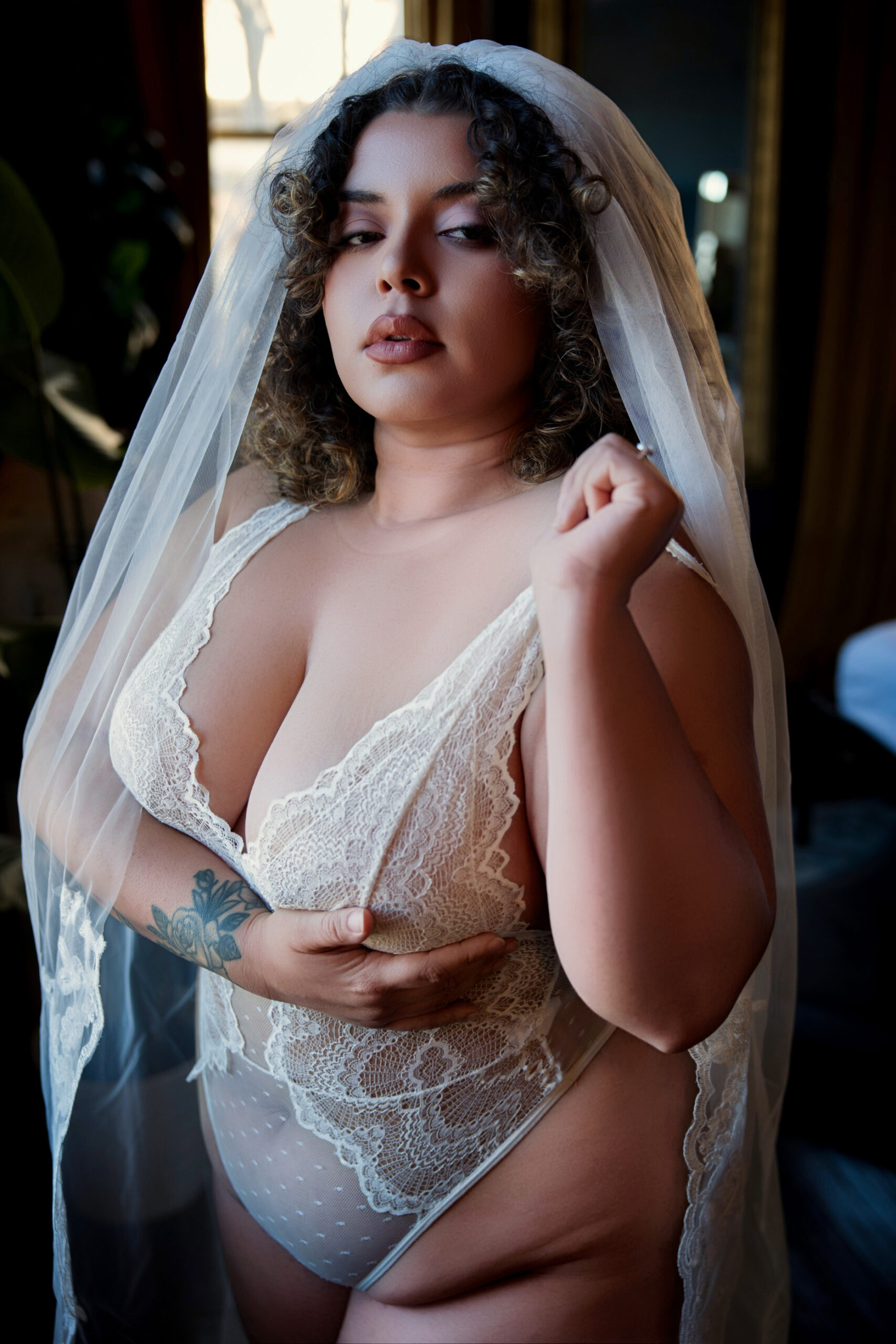 A bride stands in white lingerie and a long lace veil in a window with curly hair during her bachelorette party in northern virginia