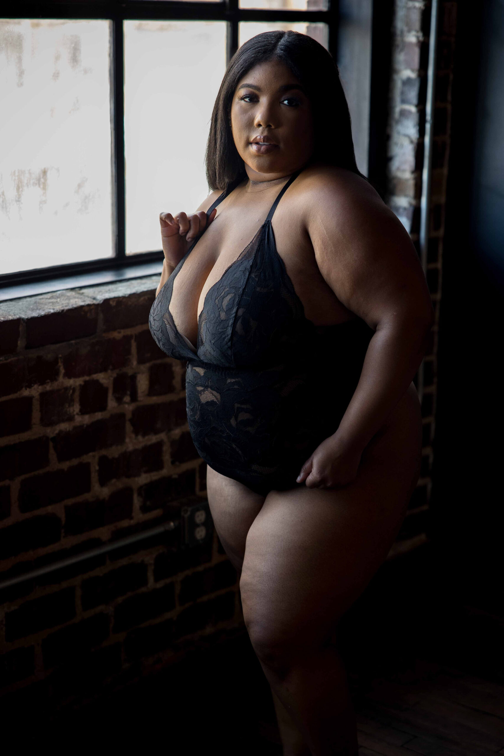 A woman stands under a window in a brick room in black lace lingerie one piece after some barre classes in baltimore
