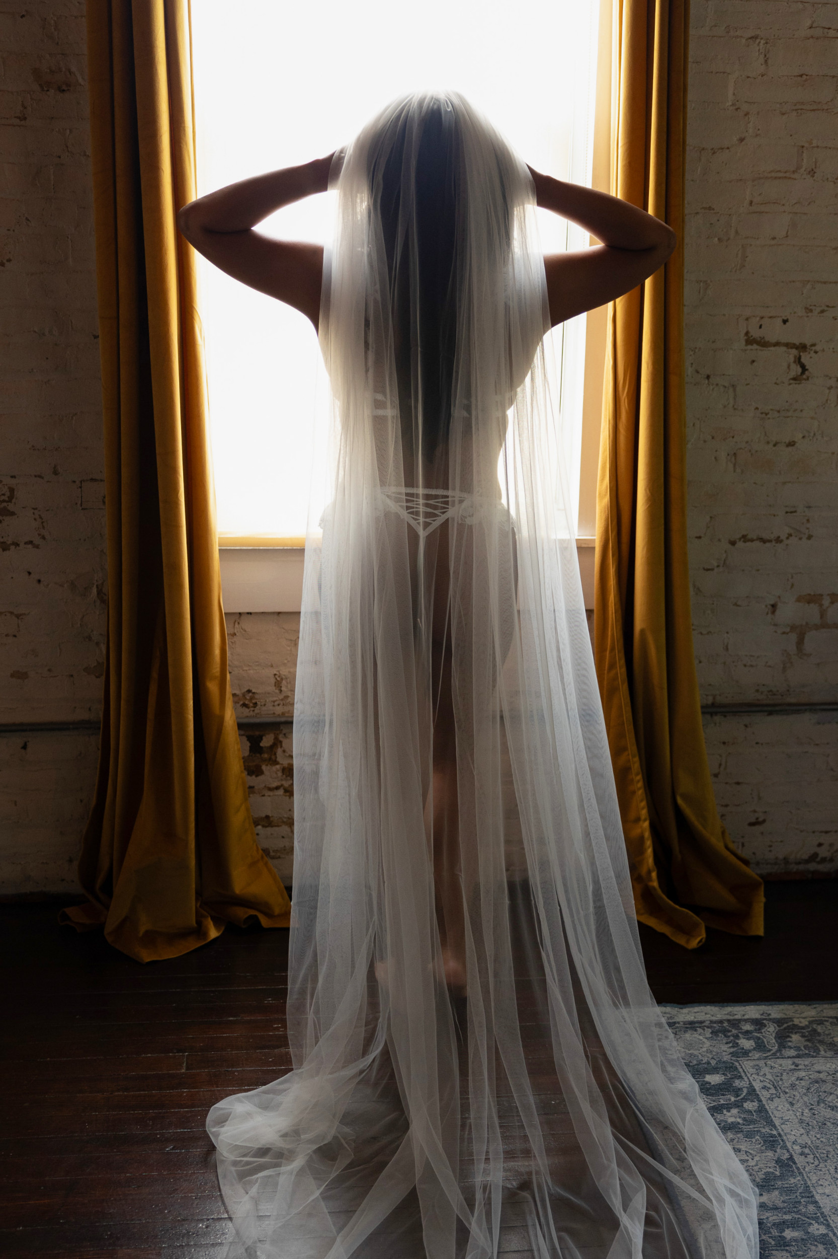 A bride stands in a window pulling back her veil while wearing white lingerie after visiting bridal shops in baltimore