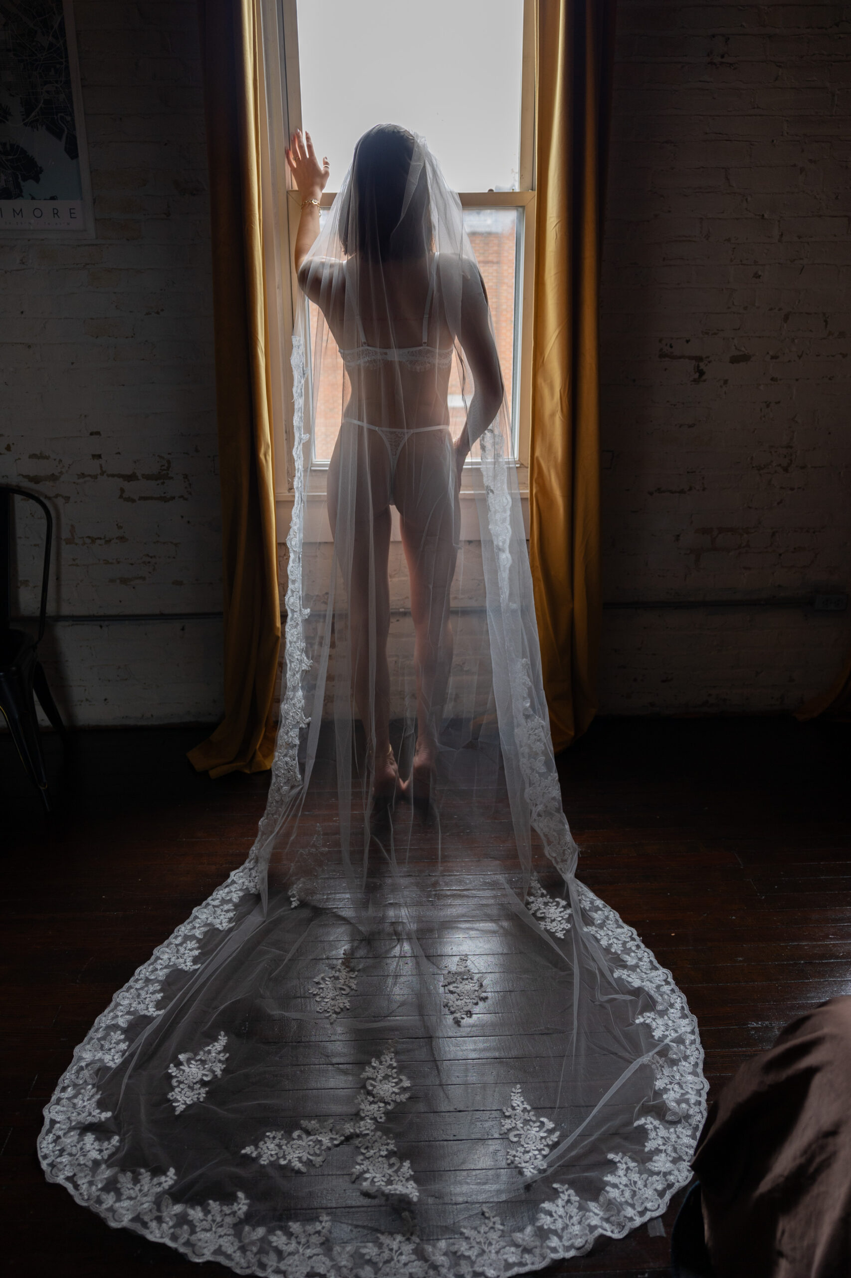 A bride stands in a window in her underwear and long lace embroidered veil spread behind her