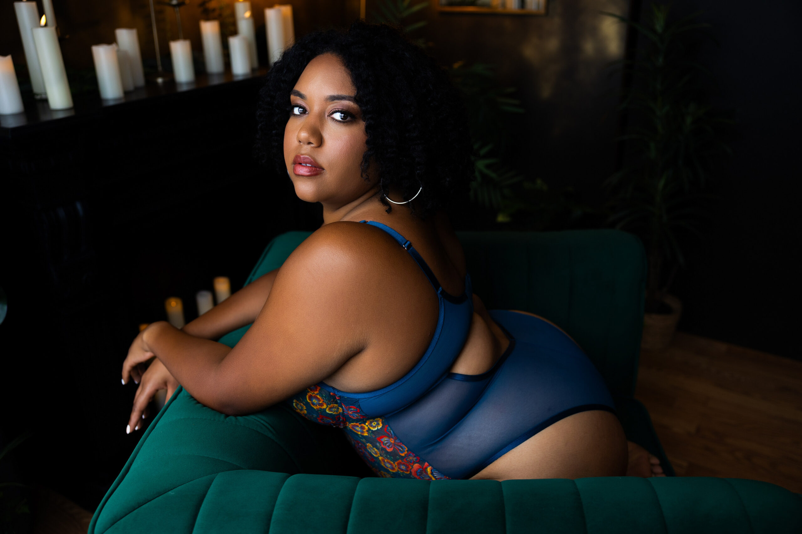 A woman in blue one piece lingerie kneels on a couch in a studio with candles looking back over her shoulder after finding lash extensions in northern virginia
