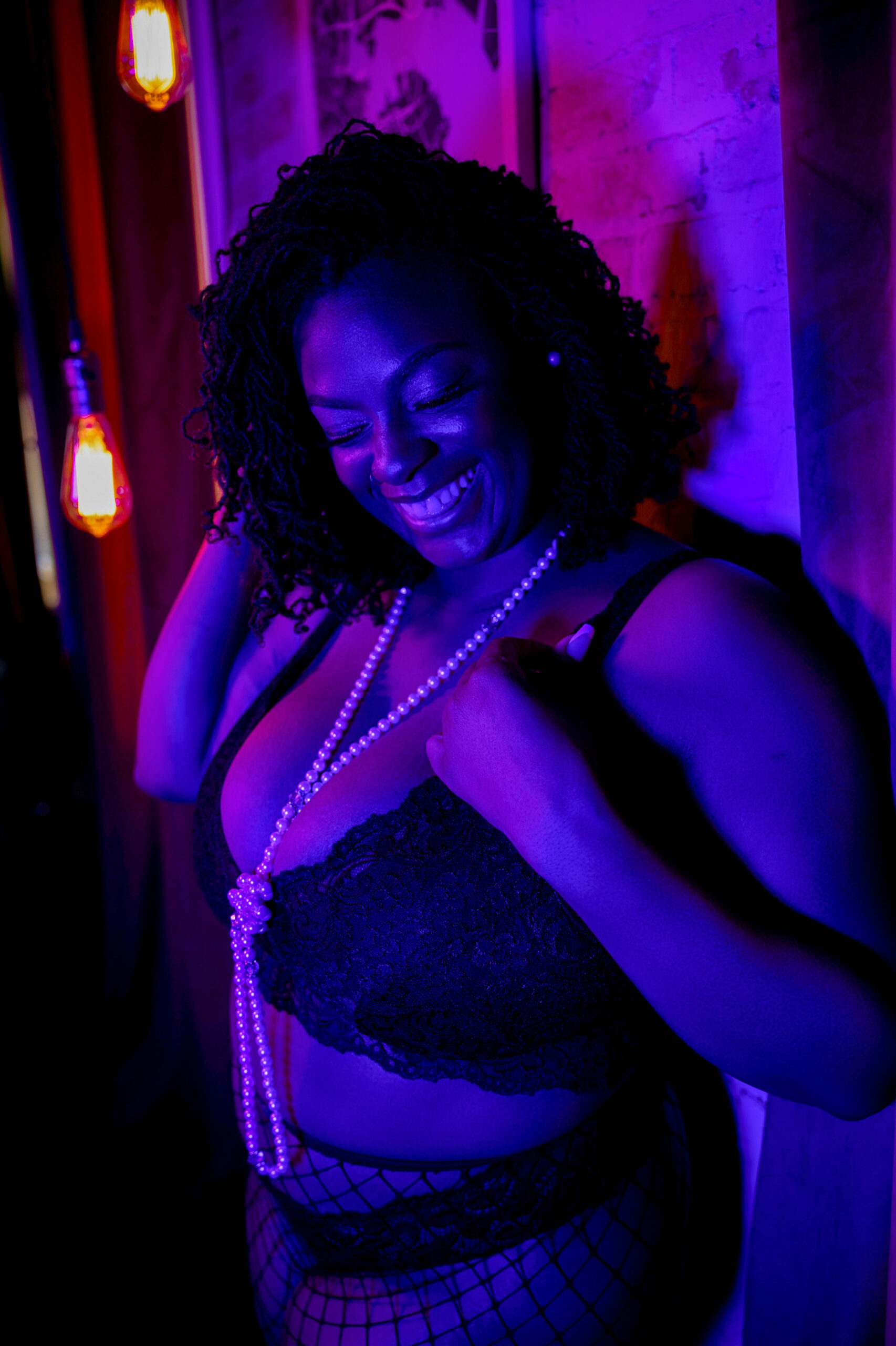 a woman in black lace bra leans against a wall smiling in long pearl necklace after finding pole fitness in baltimore