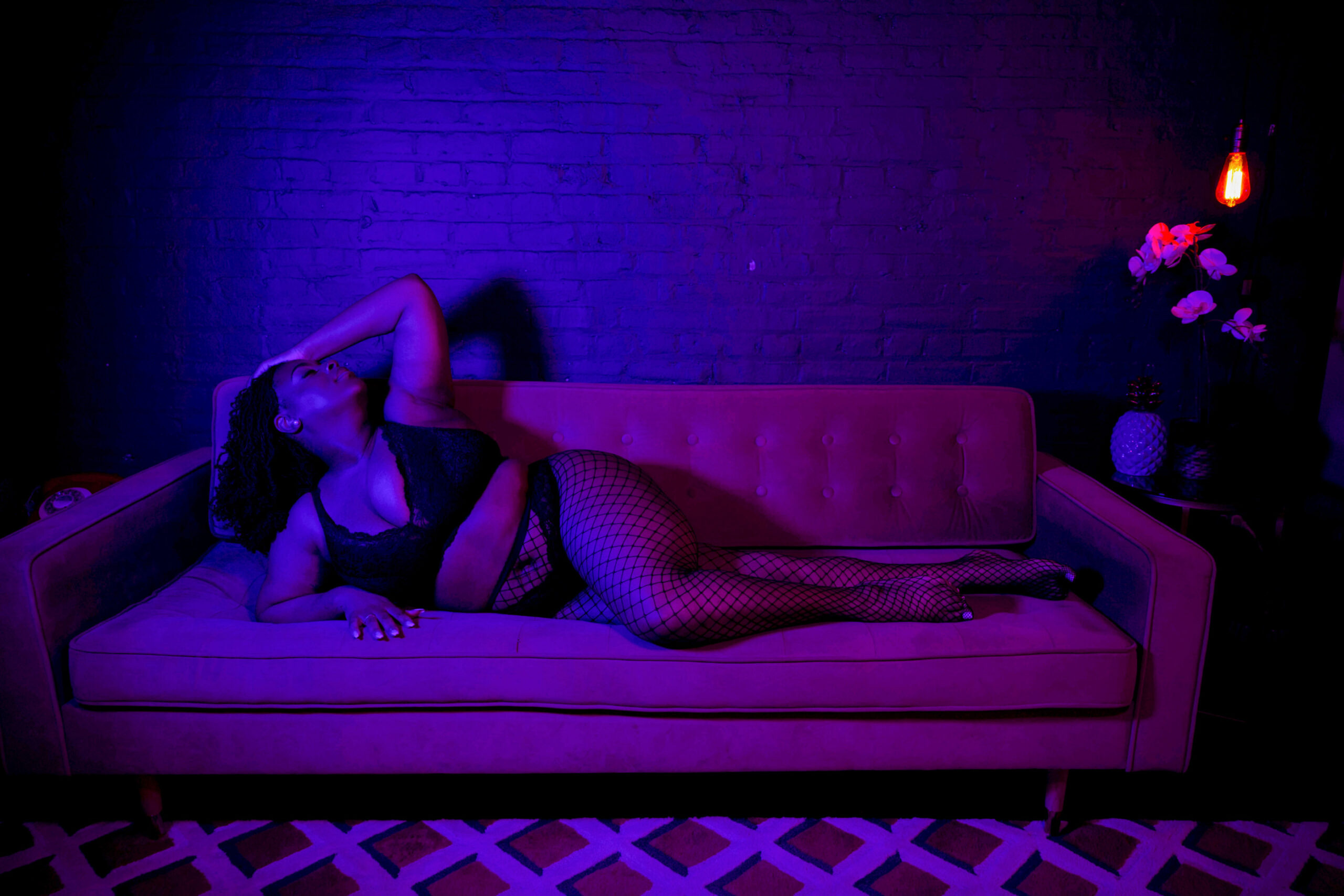 A woman in a blue and pink lit room lays across a couch in fishnets and black bra after some pole fitness in baltimore