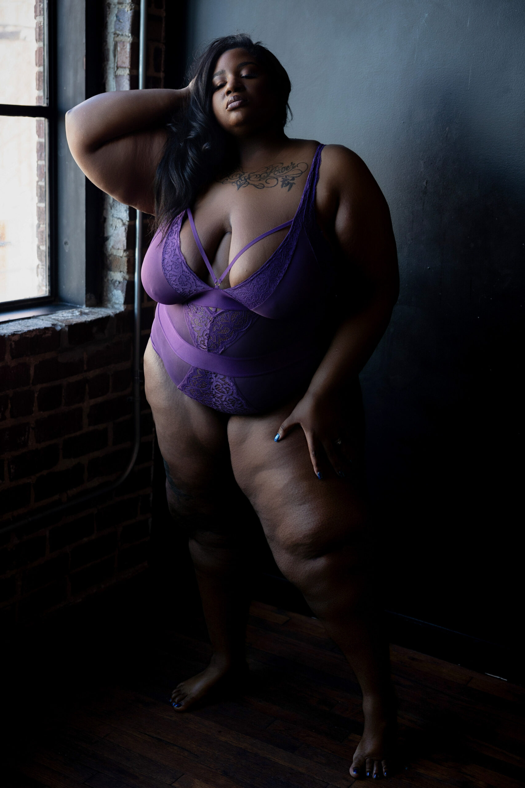A woman stands in a window in a purple lingerie body suit with a hand in her hair and one on her thigh