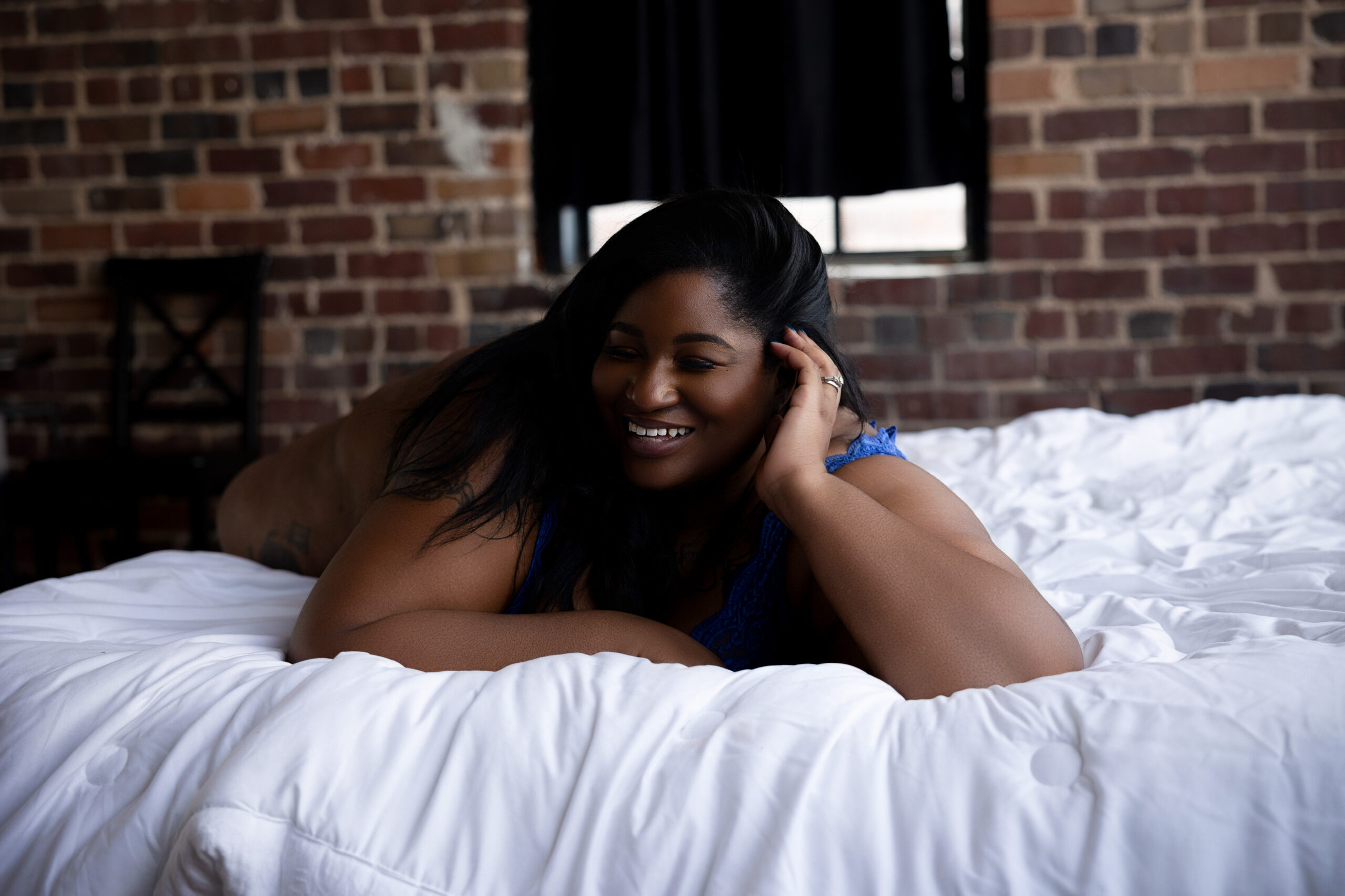 A woman laughs while laying across a white bed in a brick room in a blue bra