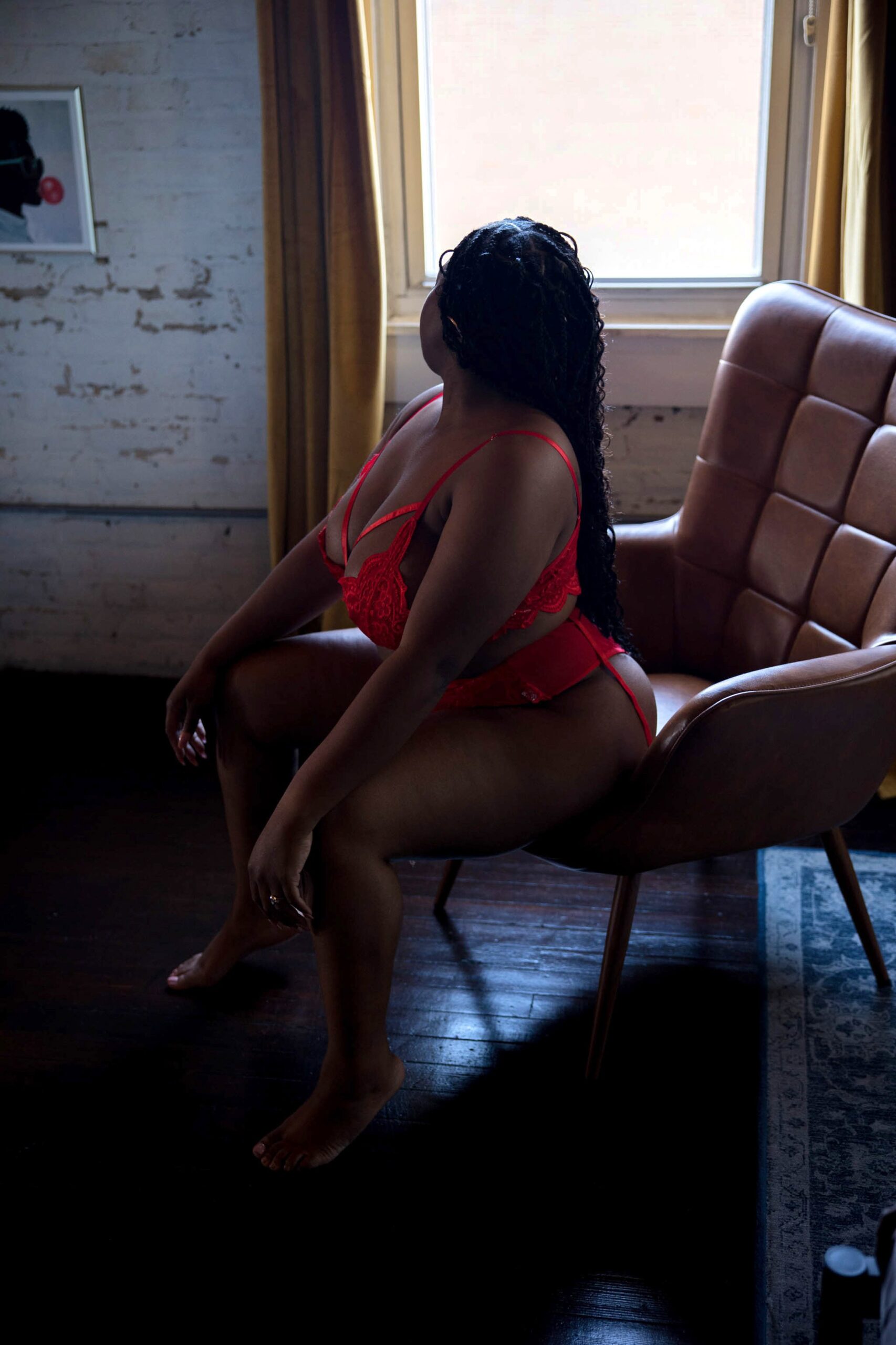 A woman looks over her shoulder while sitting on a leather chair under a window in red lingerie after visiting Renascance dermatology