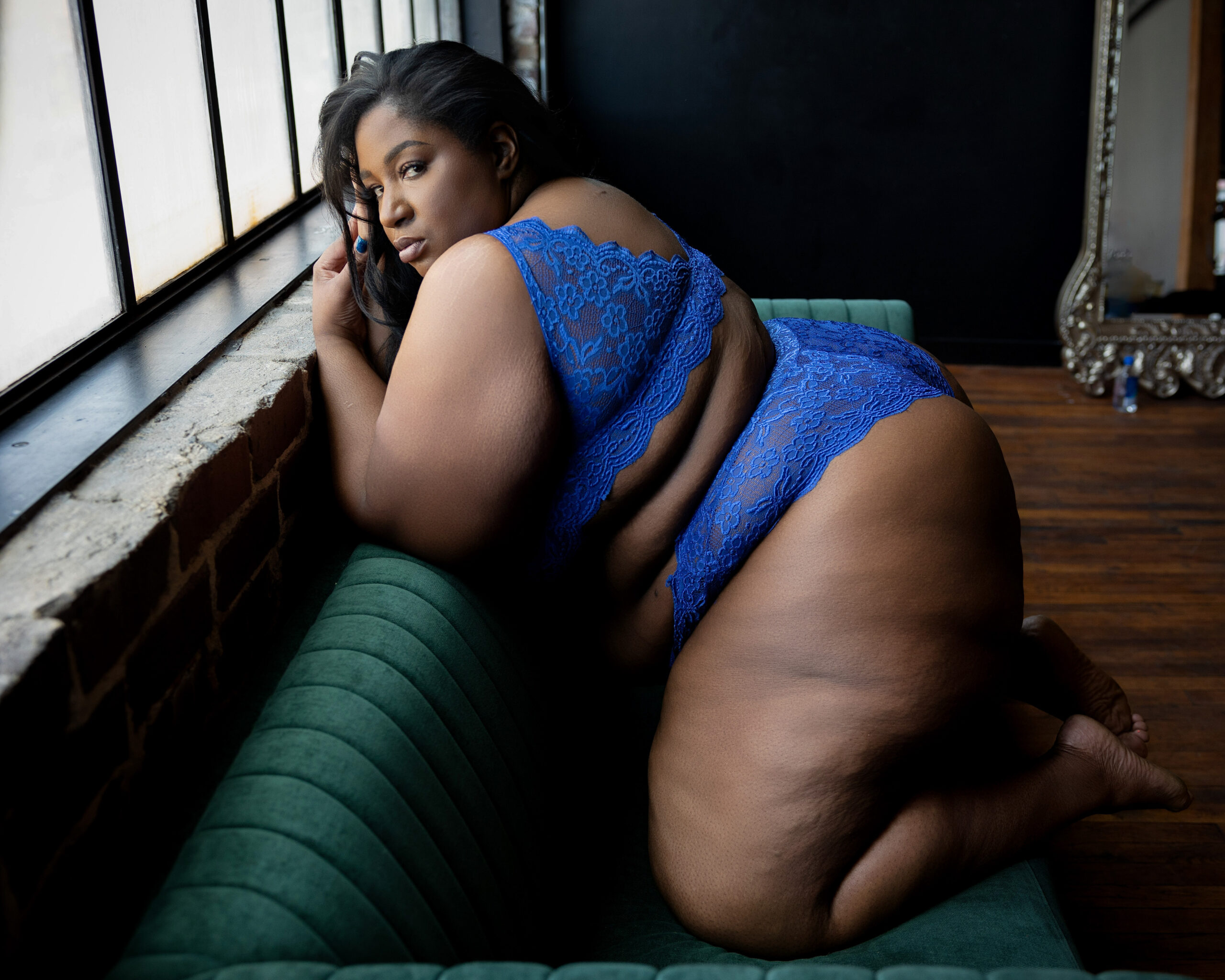 A woman in blue lace lingerie kneels on a green couch looking back over her shoulder under a window
