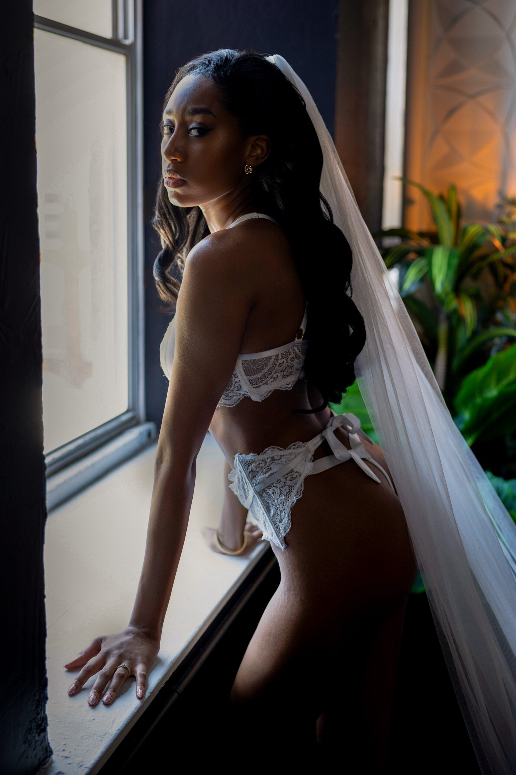 A bride wears lace bridal lingerie and a veil while leaning in a window