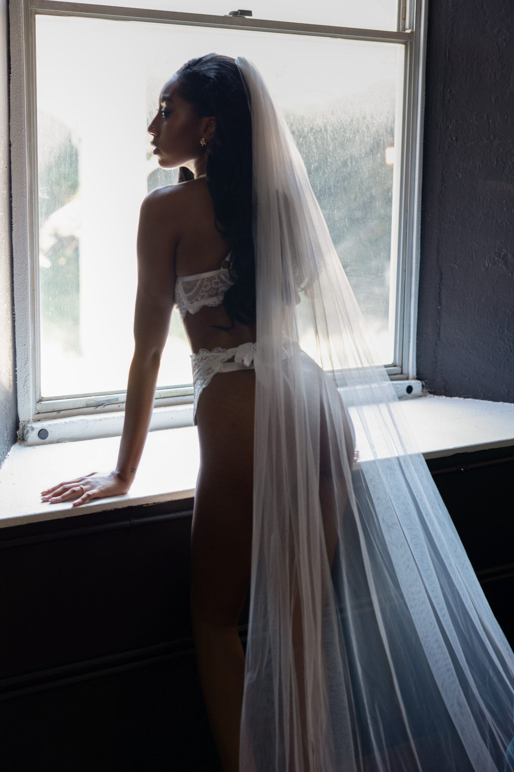 A bride leans in a window in her bridal lingerie and veil gazing over her shoulder