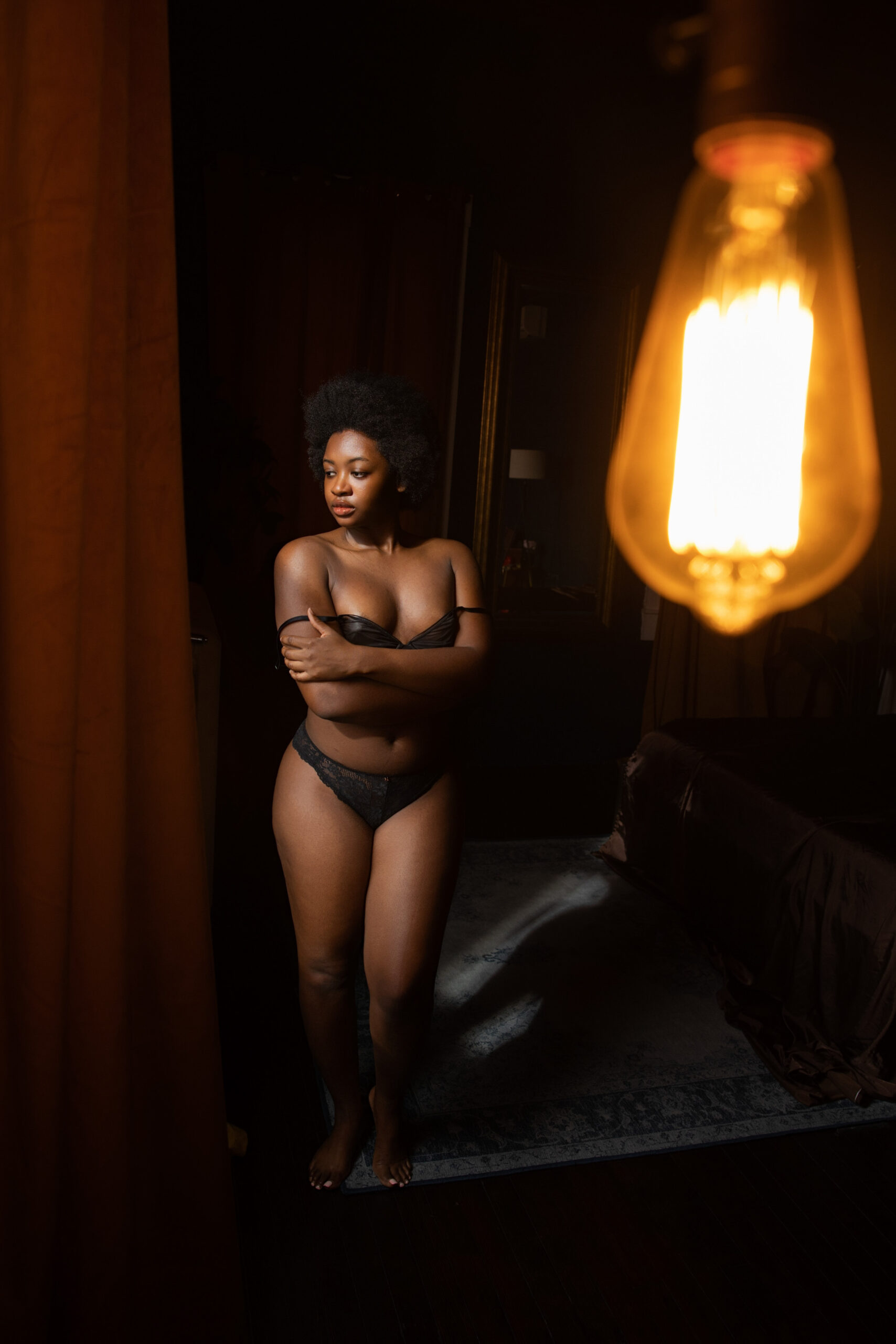 A woman covers with her arms while standing in a mirror in black underwear under a vintage lightbulb after finding medspa for skin of color northern virginia