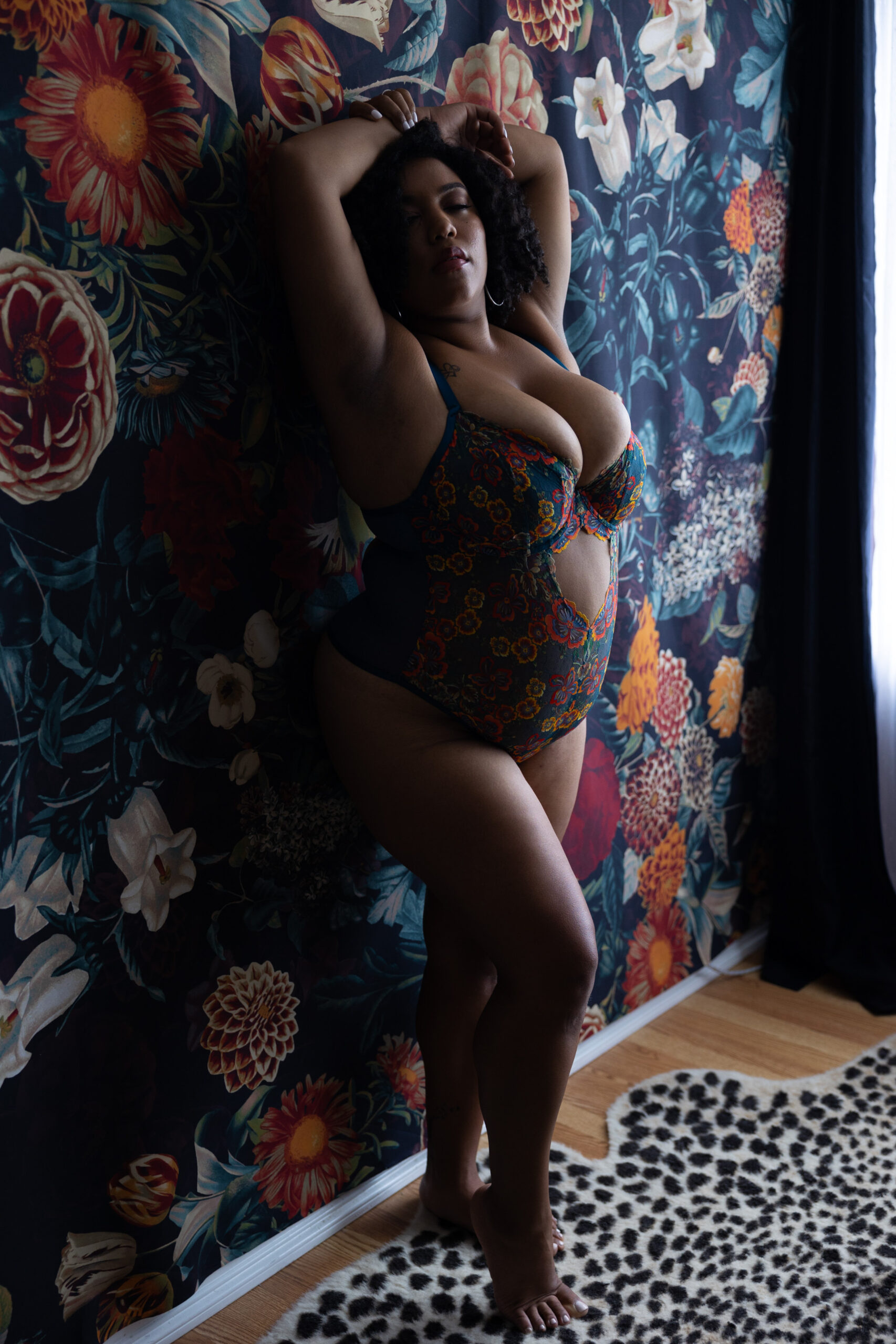 A woman in floral print bodysuit lingerie leans against a wall with hands over her head after finding waxing in dc