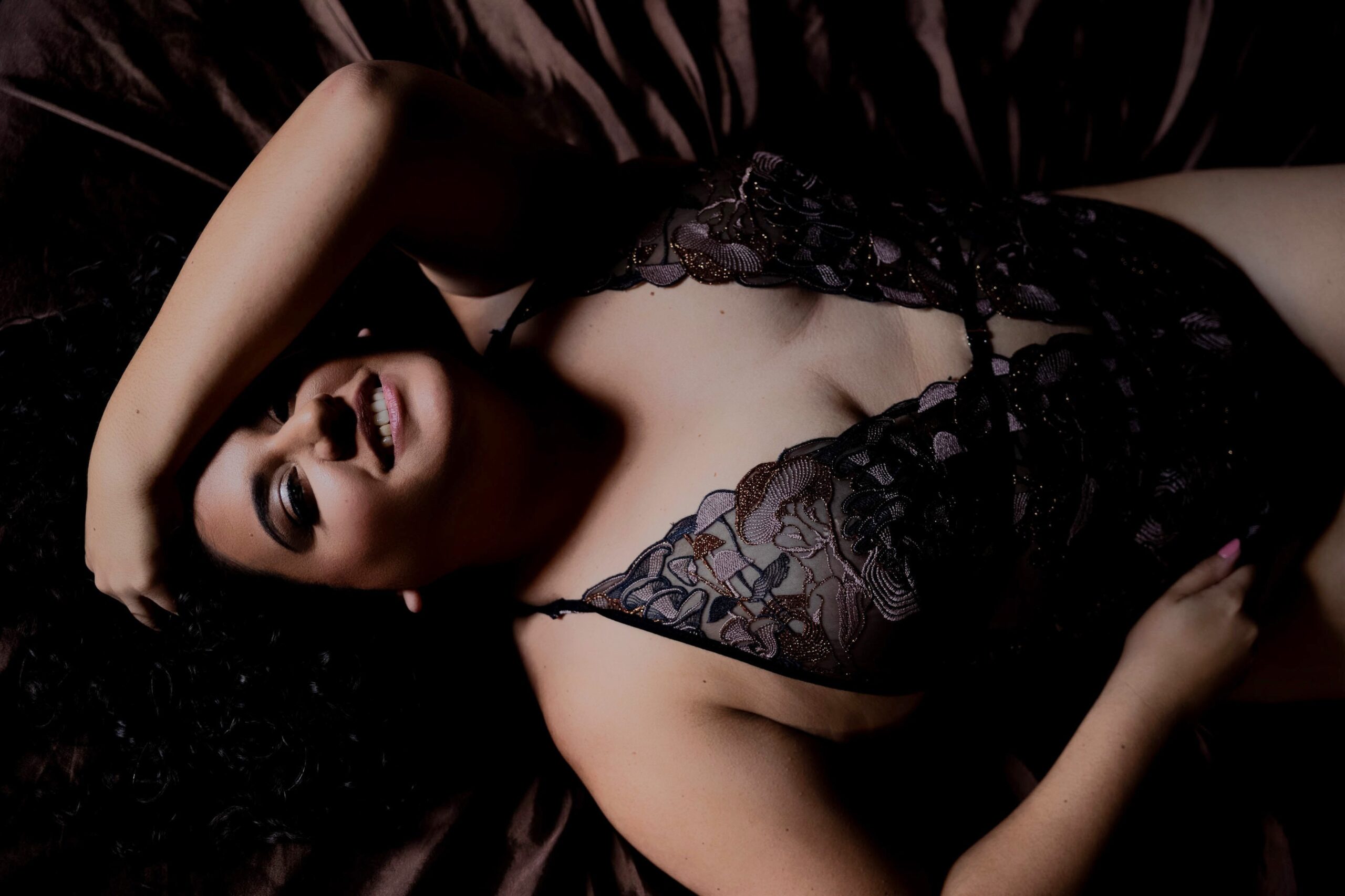 A woman smiles while laying on a bed in lace lingerie with a hand in her hair after visiting barre studios in maryland