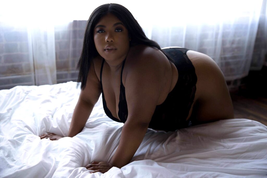 A woman bends over the edge of a bed in black lingerie under a window