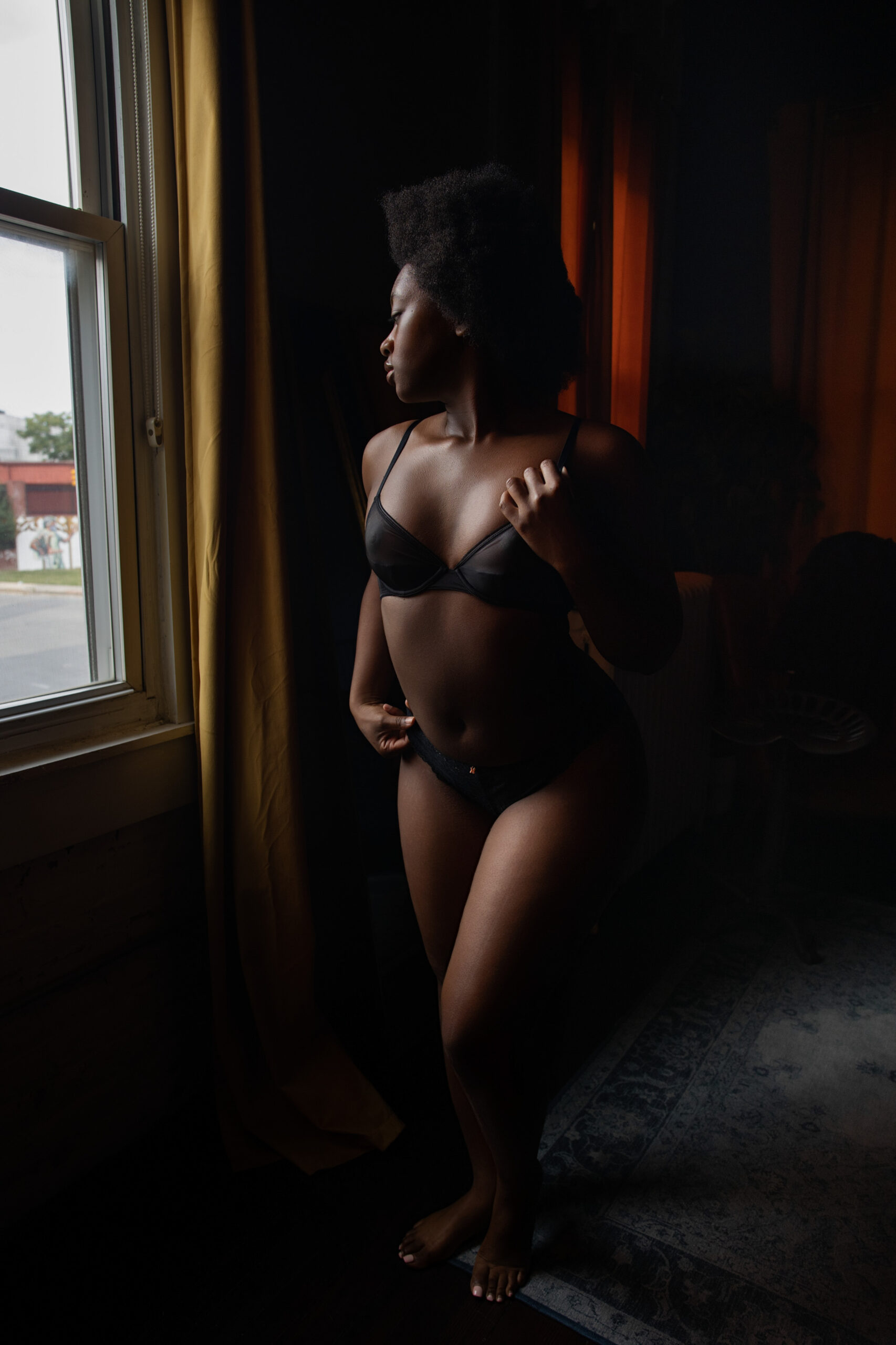 A woman stands gazing out a window while wearing black bra from a la mode intimates