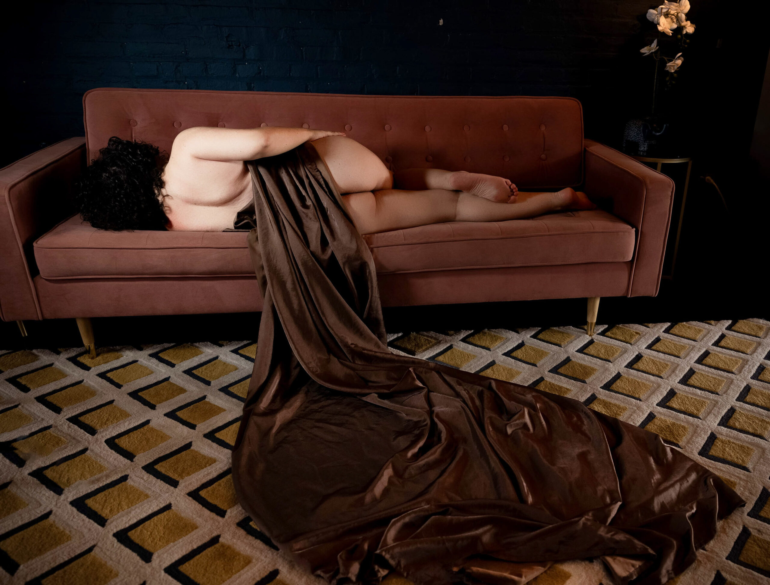A woman lays across a couch as the brown sheet covering her slips off